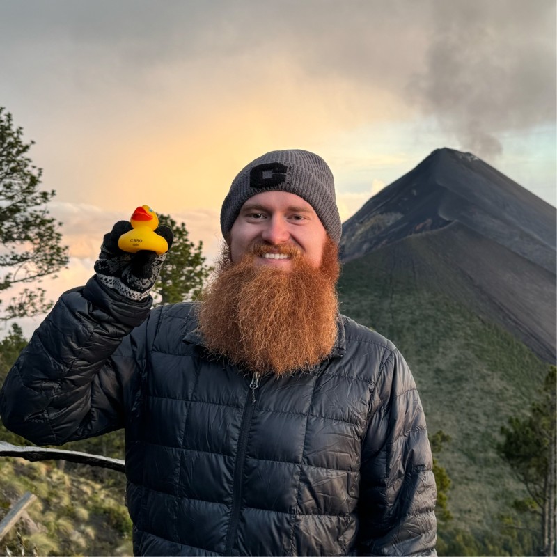 Thomas Scott Meade II in Guatemala, in front of Volcán de Fuego, wearing a well-traveled jacket and holding Patito, a small rubber duck